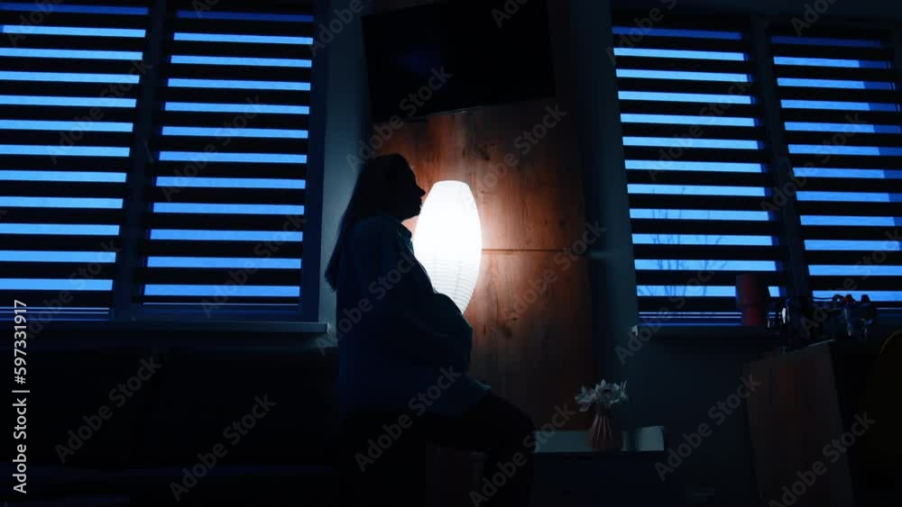 Calm long-haired pregnant woman sits on a sofa handrail in dark room. Future mom strokes her belly with love. Low angle view. Calm long-haired pregnant woman sits on a sofa handrail in dark room. Future mom strokes her belly with love. Low angle view.