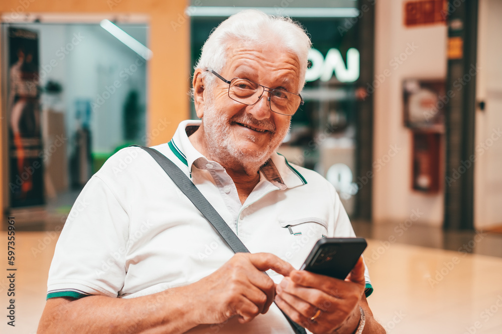 Happy older man sitting on shopping mall using smartphone. Pleasant 70s grandfather reading sms with good news, typing message, shopping in mobile app, watching photos. Happy older man sitting on shopping mall using smartphone. Pleasant 70s grandfather reading sms with good news, typing message, shopping in mobile app, watching photos.
