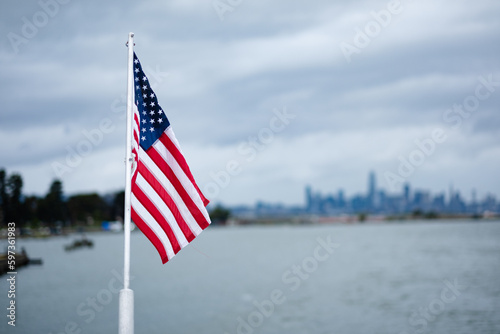 On a cloudy day, a view of the American flag on the back of the ferry from San Francisco to Oakland. They Bay Area and the San Francisco skyline are visible in the background.