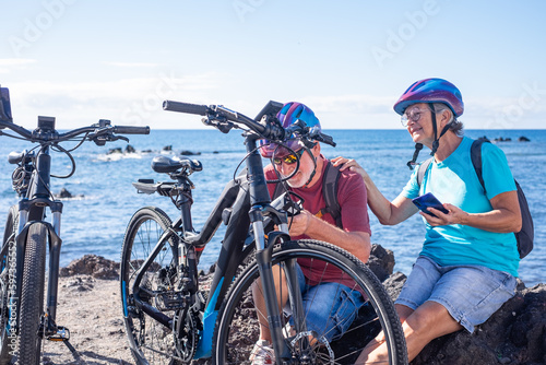 Caucasian senior cyclist couple at the beach with electric bikes in a sunny day. Old Man checks the battery while woman uses phone
