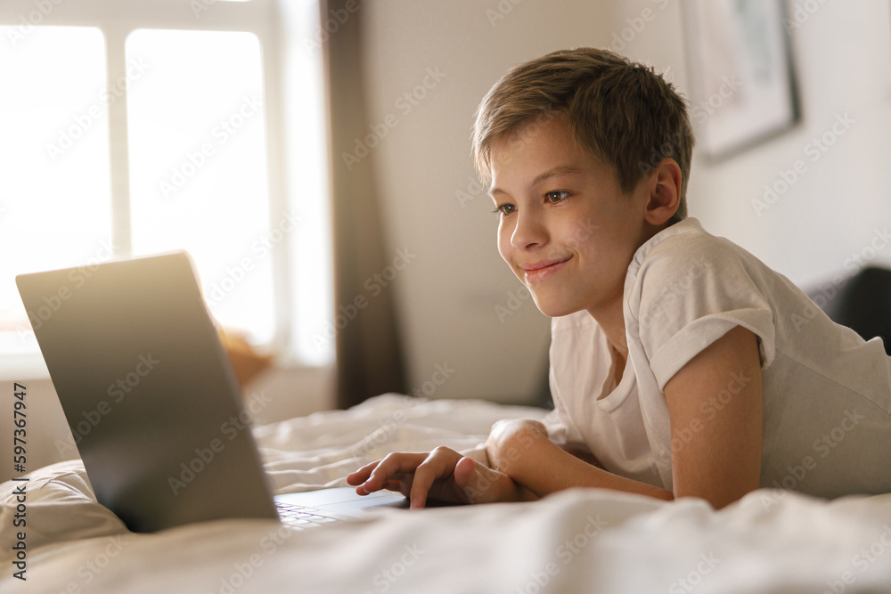 Young boy lies on the bed and looks into an open laptop. Home education concept Young boy lies on the bed and looks into an open laptop. Home education concept