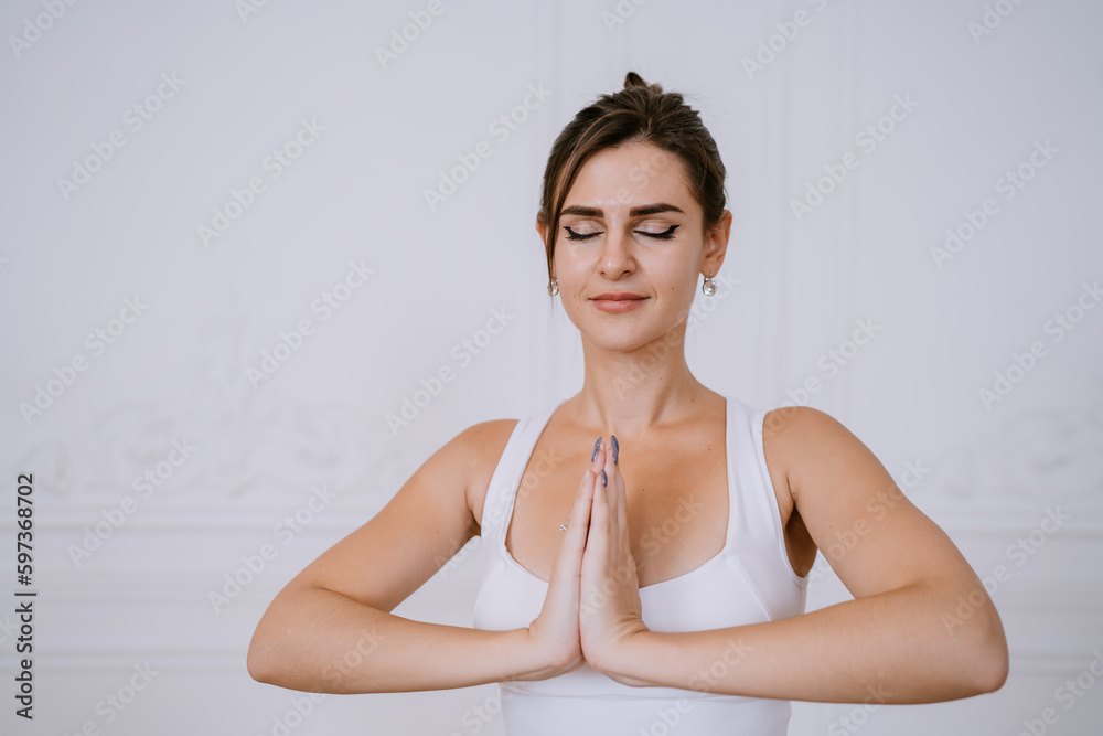Fototapeta premium Beautiful brunette American girl in sportswear meditating with hands in prayer gesture against white wall. Focused young caucasian woman concentrating on her goals preparing for yoga class.