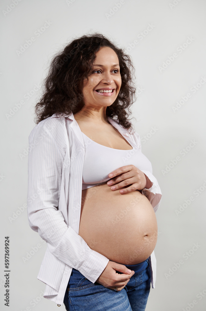 Multi ethnic curly haired gravid woman wearing blue jeans and white bra under white shirt, holds hands on her belly, smiles cutely with beautiful toothy smile, looking away, isolated white background