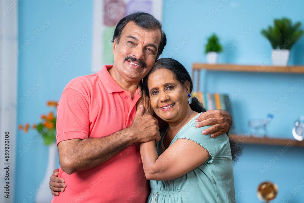 Fototapeta premium Happy Indian senior couple closely standing by embracing each other by looking camera at home - concept of family bonding, relationship and