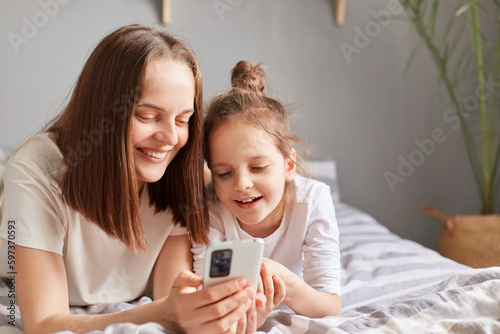 Happy mother and cute daughter using mobile phone, resting on bed at home, little girl with mommy looking at cell phone display watching video, shopping online.