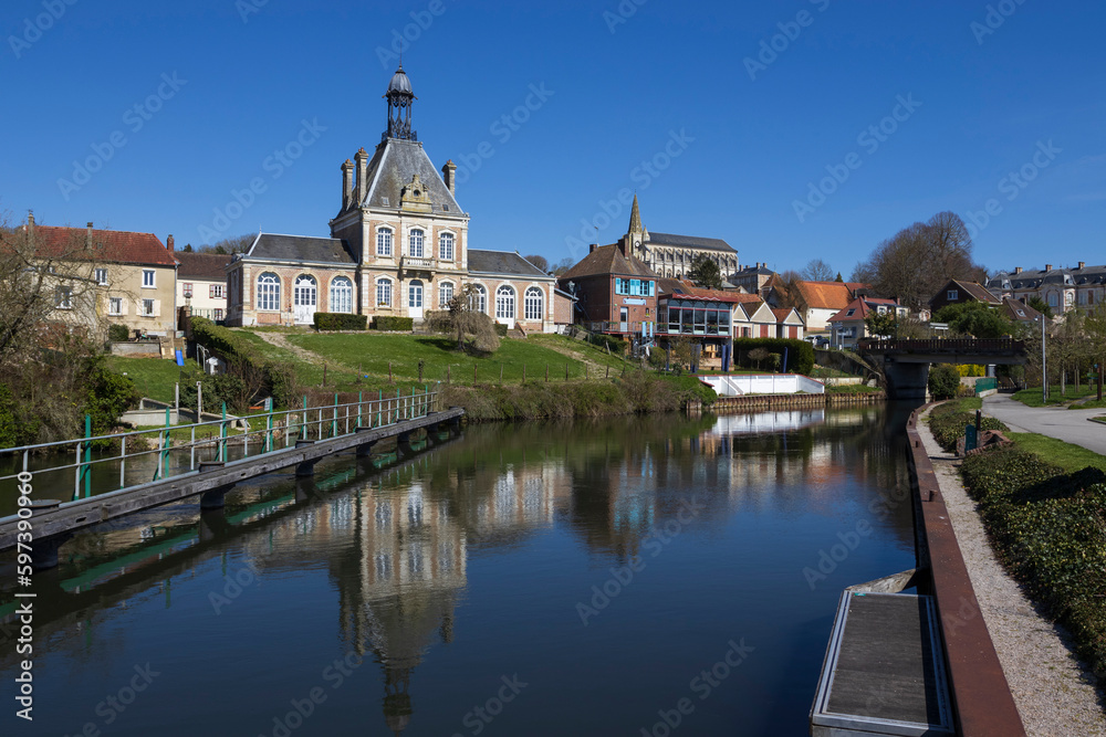 View of the River Somme and Long Town Hall in the village of Long, in the Somme department, near Abbeville in France. Sunny spring day with clear sky and reflections in the still water.