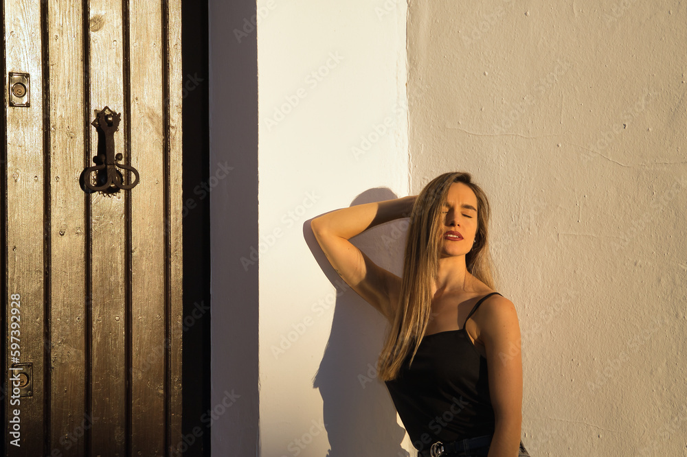 Fototapeta premium Young woman, beautiful and blonde, with black tank top receiving the last rays of afternoon sun with sensual and provocative attitude. Concept sensuality, provocation, flirting, beauty.
