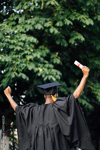 Afro american graduate from university in black mantle and hat stand with back outdoors among trees and holding higher education diploma with raised arms. Start in life, graduation, freedom.