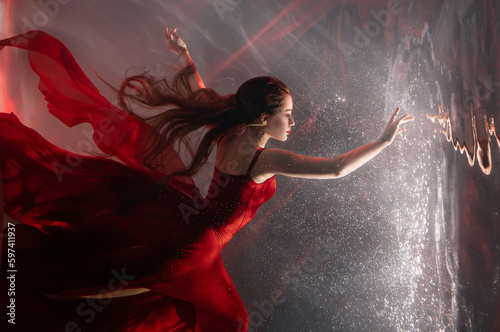 Fantasy mystery woman swims underwater touches magic light mirror, looks into reflection. Fairy tale Beauty Girl princess sleeping soars floating in dream dark water. Art ballerina dancing red dress