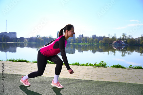 Young woman stands by the lake in the morning, dressed in sportswear, stretches her limbs, preparing to run. Her tight tank top, sleek leggings and lightweight trainers show off her toned figure.