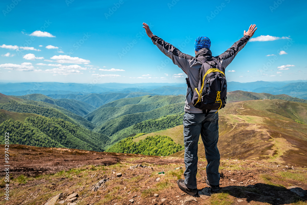 Back view, hiker with backpack standing on the mountain with raised hands up and enjoying the view
