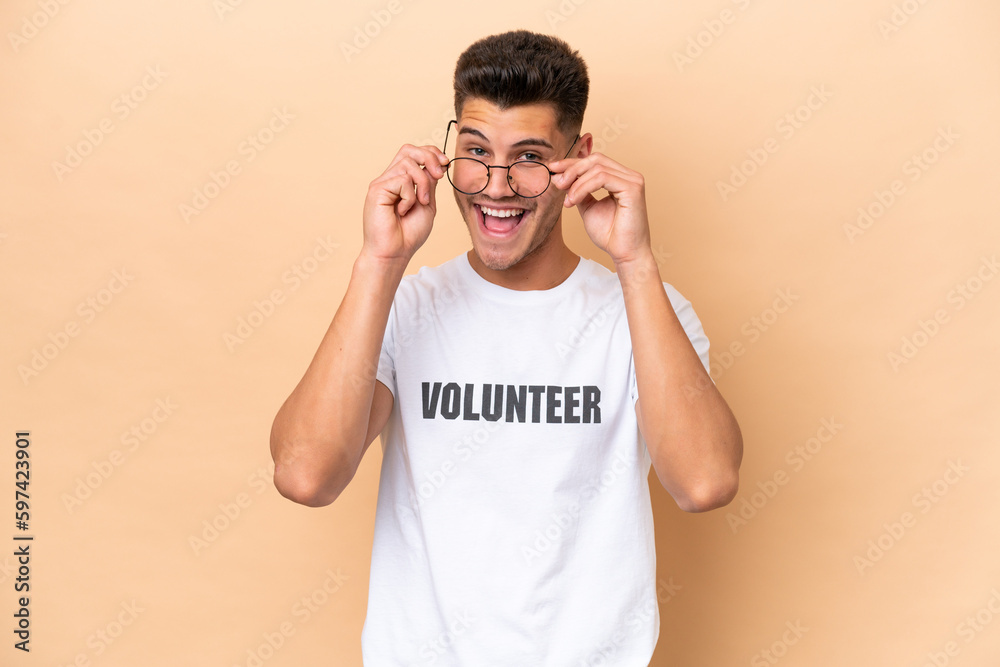 Young volunteer caucasian man isolated on beige background with glasses and surprised Young volunteer caucasian man isolated on beige background with glasses and surprised