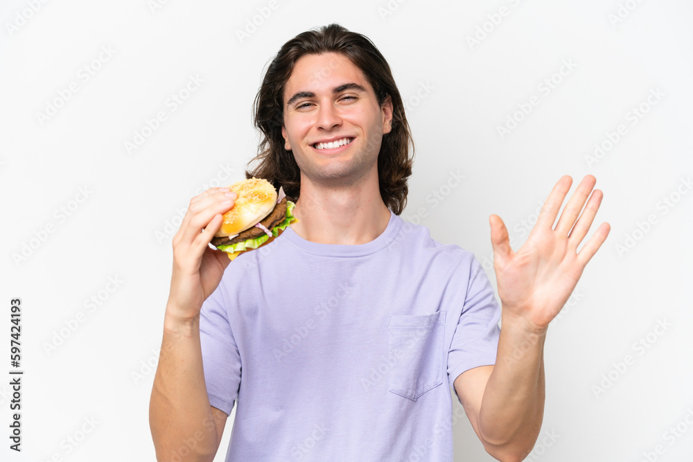 Young handsome man holding a burger isolated on white background saluting with hand with happy expression Young handsome man holding a burger isolated on white background saluting with hand with happy expression