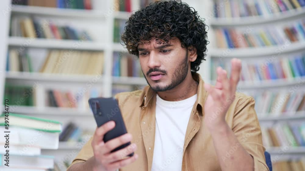 Shocked sad university student reading bad news on smartphone while sitting in campus library space. A disappointed male is upset by a text message received by email or information on the Internet