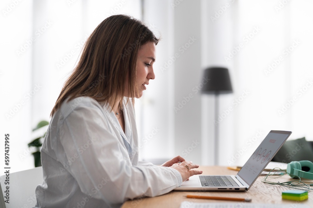 Side view of young female freelancer in casual clothes sitting at table with laptop and working on remote project Side view of young female freelancer in casual clothes sitting at table with laptop and working on remote project