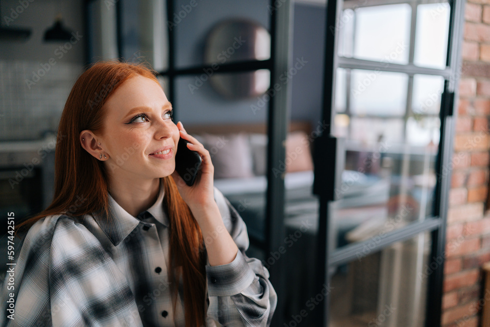 Close-up face of cheerful young woman holding mobile phone happily calling and chatting at home, smiling looking away. Closeup of laughing redhead female having pleasant conversation on smartphone. Close-up face of cheerful young woman holding mobile phone happily calling and chatting at home, smiling looking away. Closeup of laughing redhead female having pleasant conversation on smartphone.