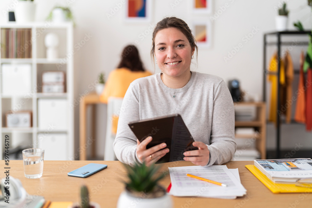Positive woman in casual clothes smiling and looking at camera while sitting at table with tablet and working in modern workspace Positive woman in casual clothes smiling and looking at camera while sitting at table with tablet and working in modern workspace