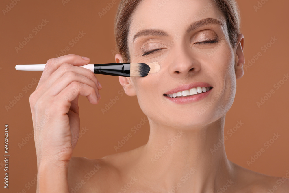Woman applying foundation on face with brush against brown background Woman applying foundation on face with brush against brown background