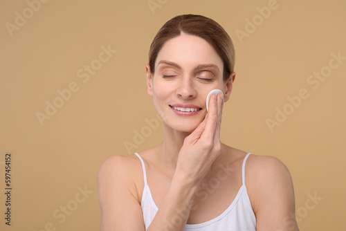 Beautiful woman removing makeup with cotton pad on beige background