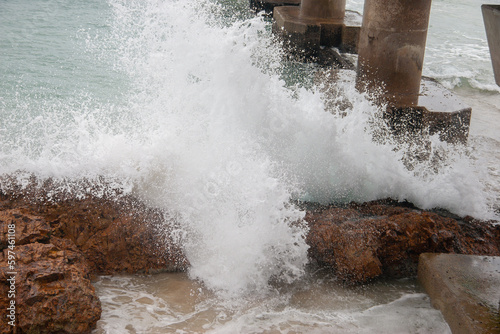 Water sprays when wave hits rugged brown rocks on seashore below pier photo