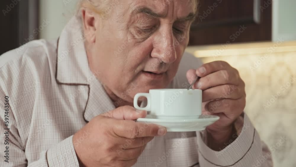 Tilt up shot of cheerful senior man in pajamas standing in kitchen at home, stirring freshly made coffee, enjoying smell and smiling Tilt up shot of cheerful senior man in pajamas standing in kitchen at home, stirring freshly made coffee, enjoying smell and smiling