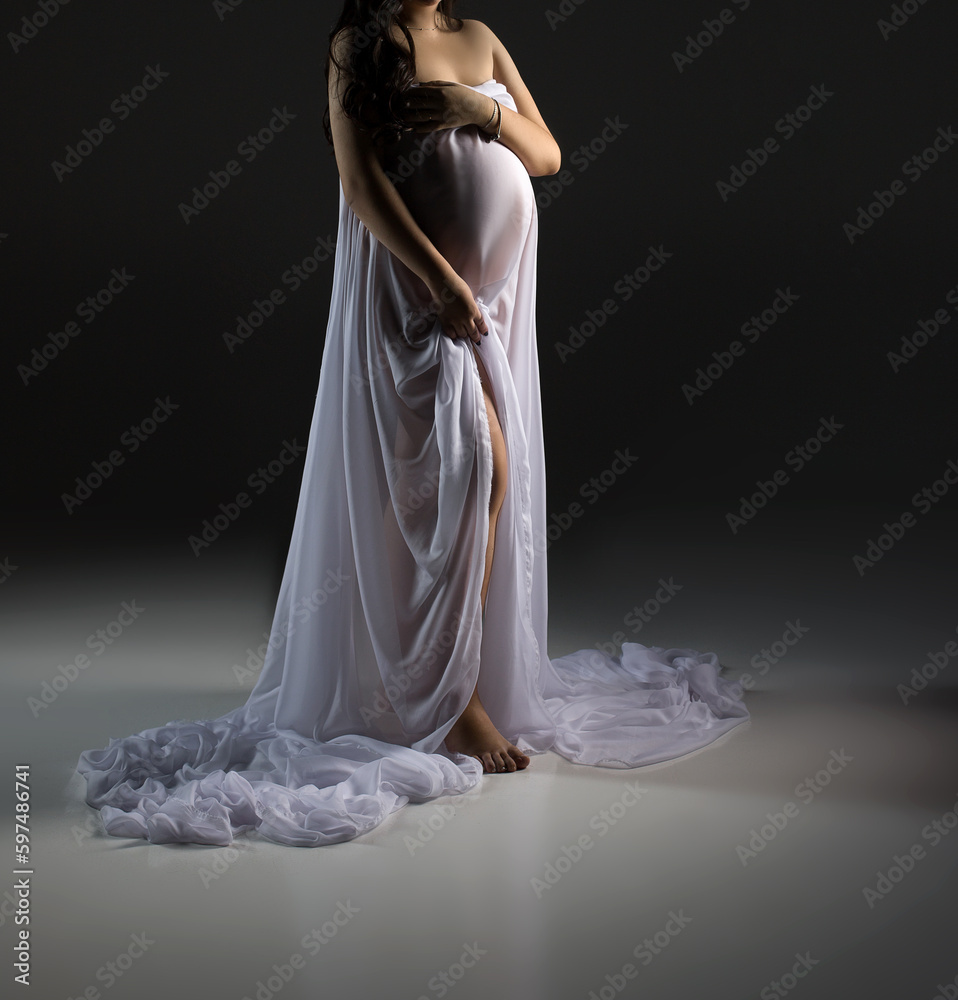 A pregnant woman is standing in a white dress on a black background. Studio pregnancy photo shoot. Space for text. Woman in white dress. A pregnant woman is standing in a white dress on a black background. Studio pregnancy photo shoot. Space for text. Woman in white dress.