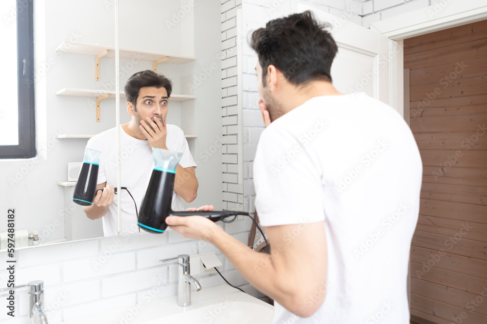 Funny man standing in front of the mirror and amusing with hair dryer in the bathroom during daily morning routine. Bad hair day concept Funny man standing in front of the mirror and amusing with hair dryer in the bathroom during daily morning routine. Bad hair day concept