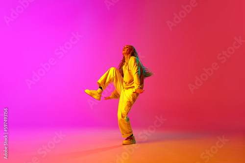 Portrait with modern young dancer wearing fashion clothes in motion over gradient pink background in neon light. Funny dance