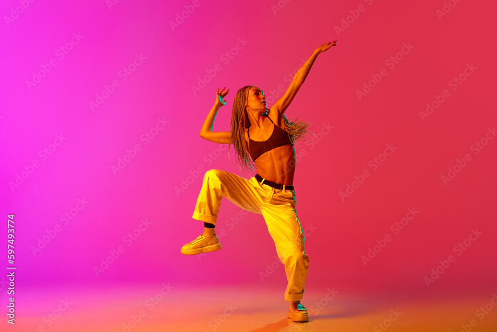 Portrait of young stylish woman, hip-hop dancer training in casual clothes over gradient pink background in neon light. Street style dance