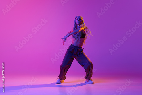 One professional hip-hop dancer wearing fashion clothes moving with inspiration over gradient purple background in neon light. Modern choreography