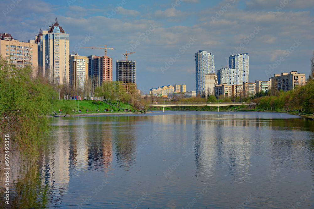 Wide-angle landscape view of water channel near Rusanivka neighborhood. High water in Kyiv, spring 2023, Ukraine. Residential high-rise buildings in the background against blue sky. Flooding