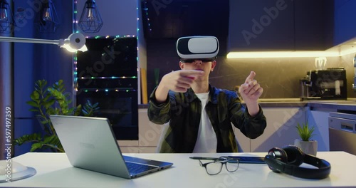 Teenager boy wearing VR glasses moving hands in air scroling typing, sitting on desk in living room. Virtual reality, modern gadget device. photo