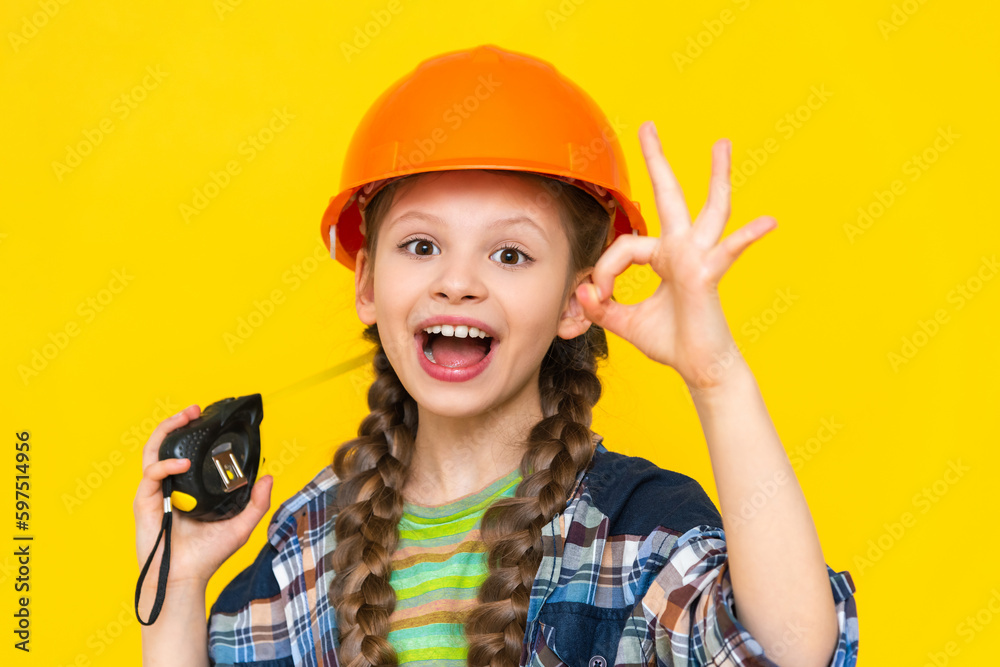 A teenage girl in a construction helmet shows an ok sign and holds a tape measure. The child is an engineer. The concept of renovation in the children's room. A teenage girl in a construction helmet shows an ok sign and holds a tape measure. The child is an engineer. The concept of renovation in the children's room.