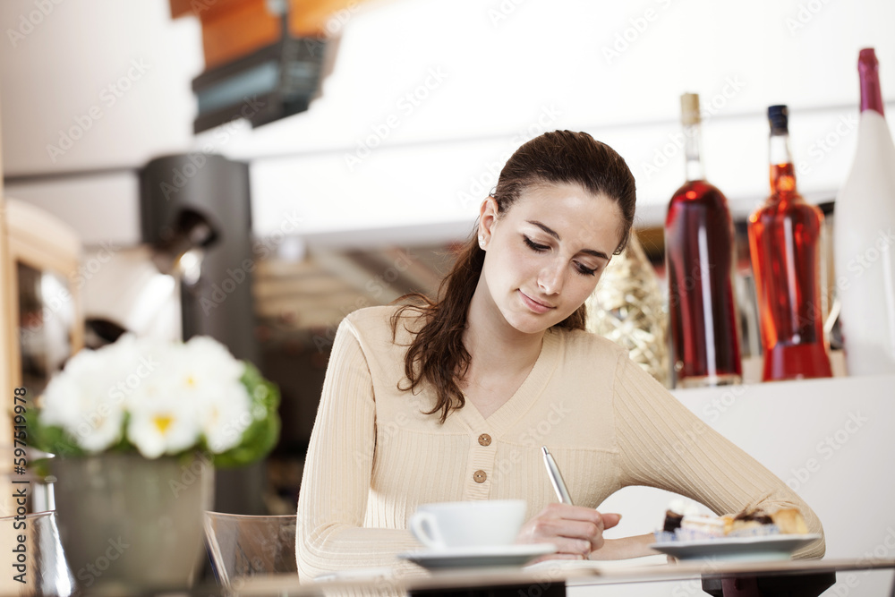 Beautiful smiling young woman writes and drinks coffee at the cafe, ice cream shop and pastry shop. Portrait of brunette student girl studying, writing on her notebook Beautiful smiling young woman writes and drinks coffee at the cafe, ice cream shop and pastry shop. Portrait of brunette student girl studying, writing on her notebook