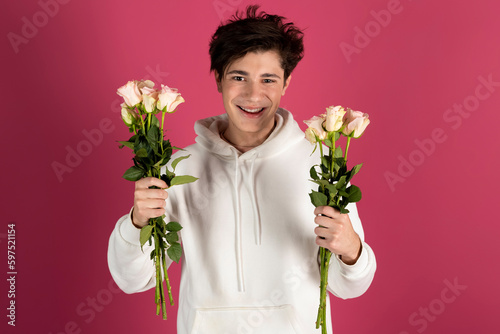Happy, handsome young man in white hoodie, holds out bouquets of flowers or roses. going to a date. Isolated on pink.
