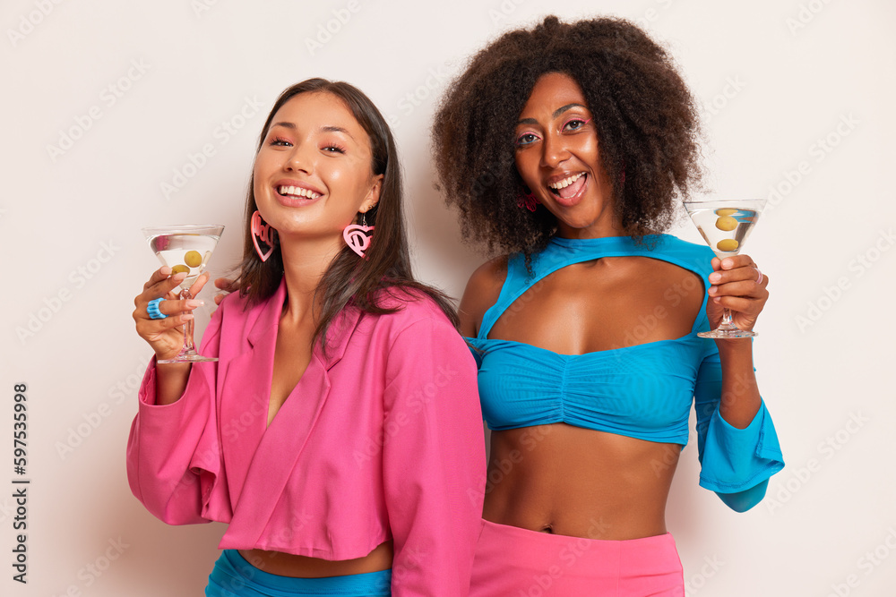 Couple of beautiful girls having fun in the studio, posing together drinking martini, wearing pink and blue clothes, happy time concept, copy space Couple of beautiful girls having fun in the studio, posing together drinking martini, wearing pink and blue clothes, happy time concept, copy space