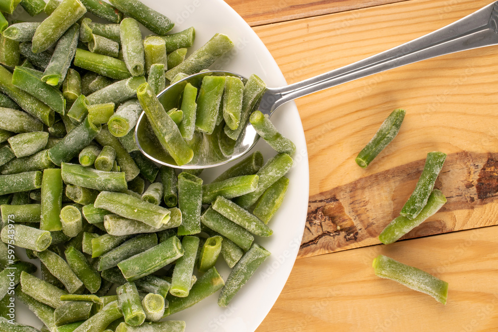 Frozen green beans in a white ceramic plate with a metal spoon on a wooden table, macro, top view. Frozen green beans in a white ceramic plate with a metal spoon on a wooden table, macro, top view.