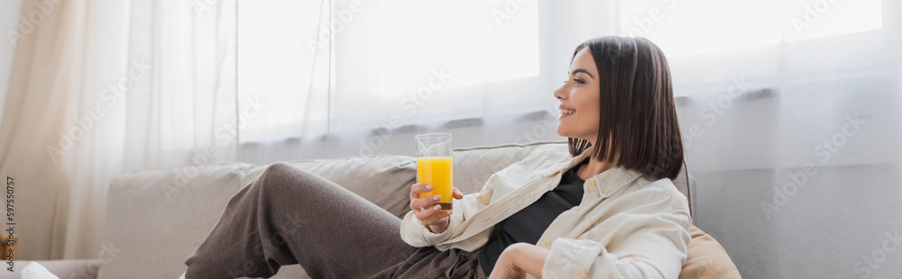 Side view of overjoyed woman holding orange juice while sitting on couch in living room, banner. Side view of overjoyed woman holding orange juice while sitting on couch in living room, banner.