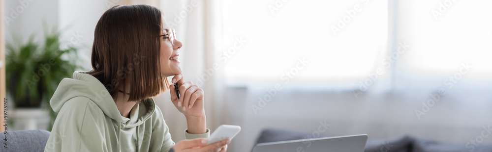 Side view of smiling freelancer in eyeglasses holding cellphone near laptop at home, banner. Side view of smiling freelancer in eyeglasses holding cellphone near laptop at home, banner.
