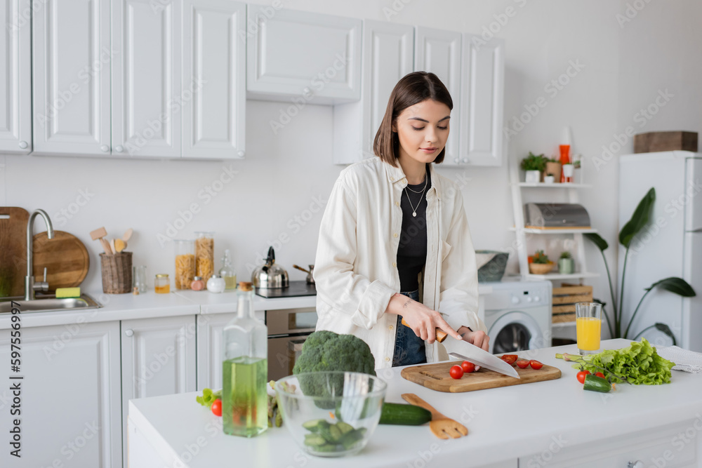 Brunette woman cutting cherry tomatoes near organic vegetables in kitchen. Brunette woman cutting cherry tomatoes near organic vegetables in kitchen.