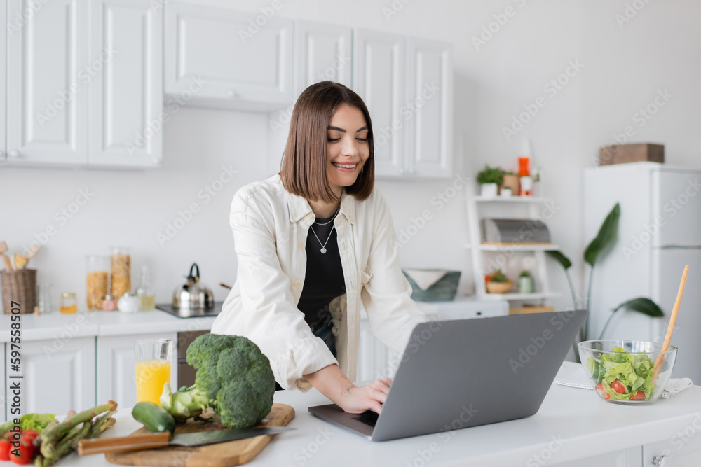 Positive woman using laptop near ripe vegetables and orange juice in kitchen. Positive woman using laptop near ripe vegetables and orange juice in kitchen.