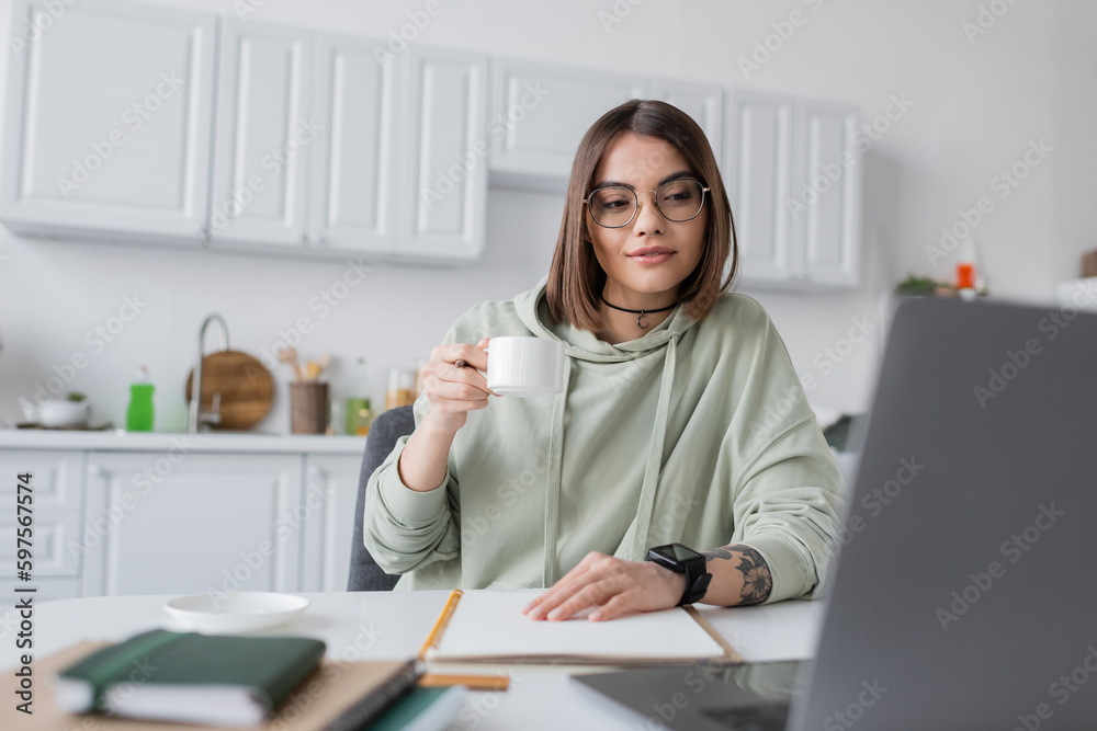 Tattooed freelancer in eyeglasses holding coffee near blurred laptop and notebooks at home. Tattooed freelancer in eyeglasses holding coffee near blurred laptop and notebooks at home.