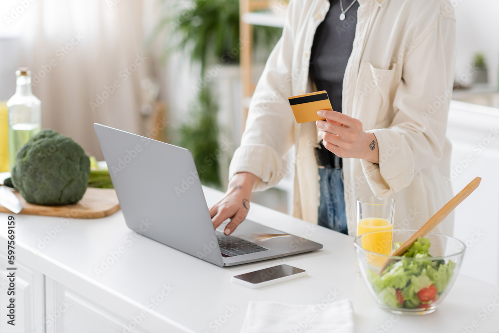Cropped view of tattooed woman holding credit card and using laptop near blurred salad in kitchen. Cropped view of tattooed woman holding credit card and using laptop near blurred salad in kitchen.