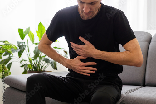 The man is sitting on a gray couch and holding his belly. Medicine and health concept, stomach problems. The man suffers from stomach ache, gastric problems. Abdominal pain, suffering and pain