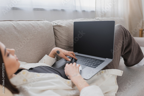 Blurred tattooed freelancer using laptop with blank screen on couch at home. © LIGHTFIELD STUDIOS