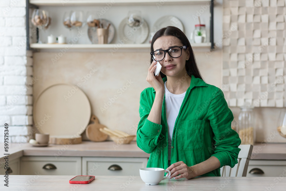 Upset young woman sitting at home in the kitchen with a mobile phone and crying. He looks sadly at the camera, wipes his tears with a napkin.