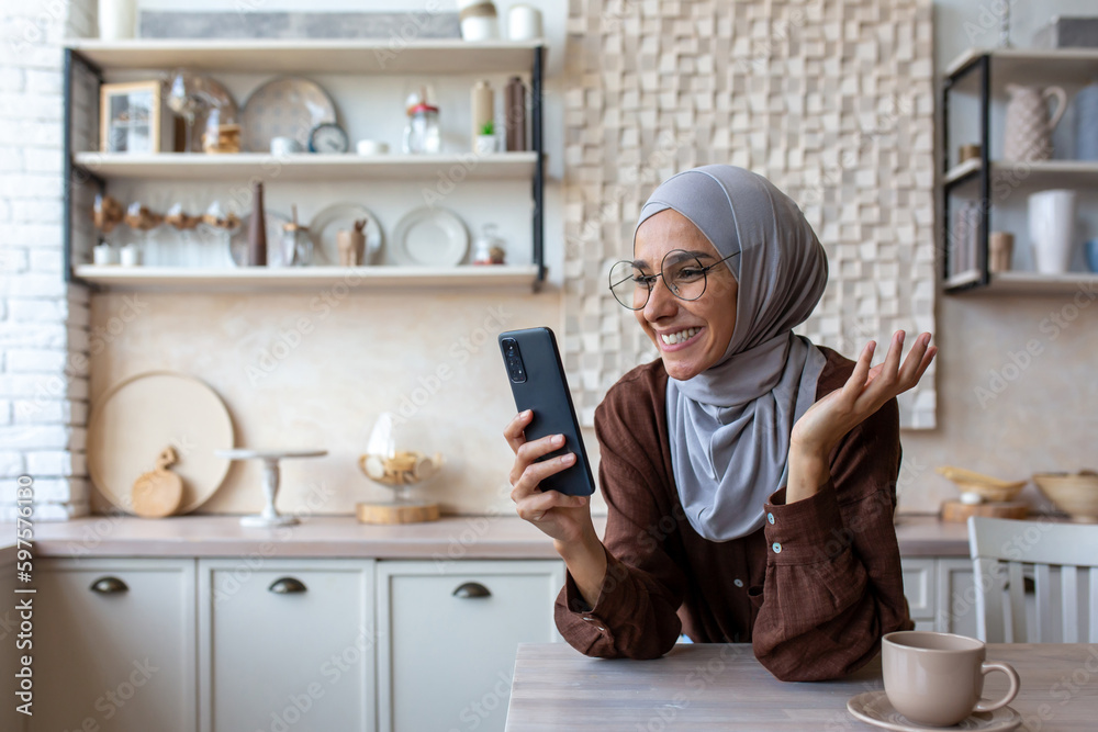 Close-up photo. Smiling Muslim young woman in hijab standing in kitchen at home, leaning on table and talking on mobile phone video call.