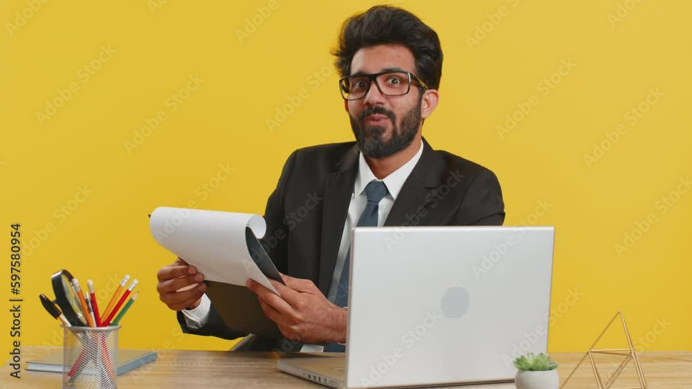 Happy indian businessman reading business contract, planning project, analyzing resume, satisfied with good paperwork results at office workplace desk. Hindu man working on laptop on yellow background Happy indian businessman reading business contract, planning project, analyzing resume, satisfied with good paperwork results at office workplace desk. Hindu man working on laptop on yellow background