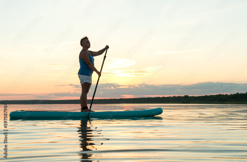 A woman in shorts and a top on a SUP board with a paddle at sunset swims in the water of the lake.