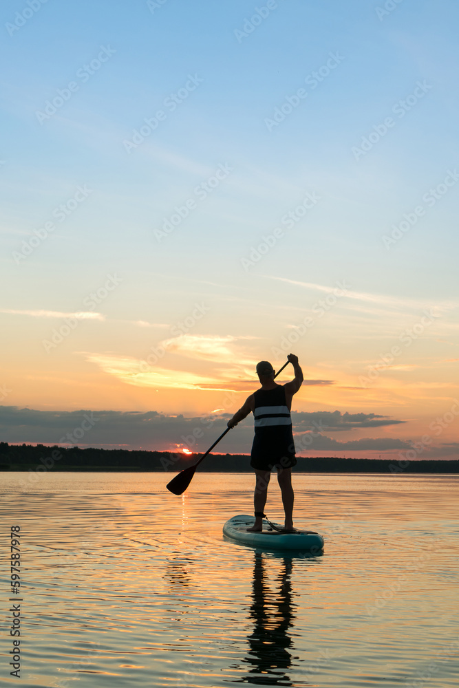 A man in shorts stands on a SUP board with a paddle at sunset in a lake against the backdrop of a sunset sky and water.